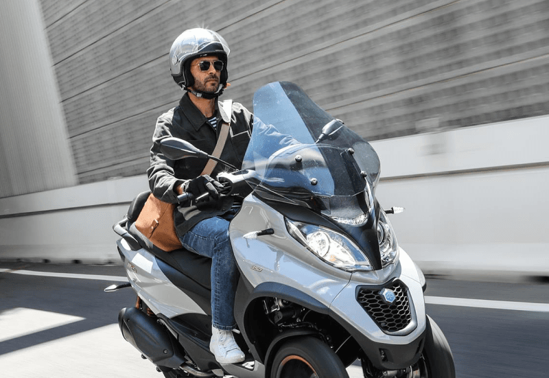 A male riding on a white three-wheeled Piaggio scooter