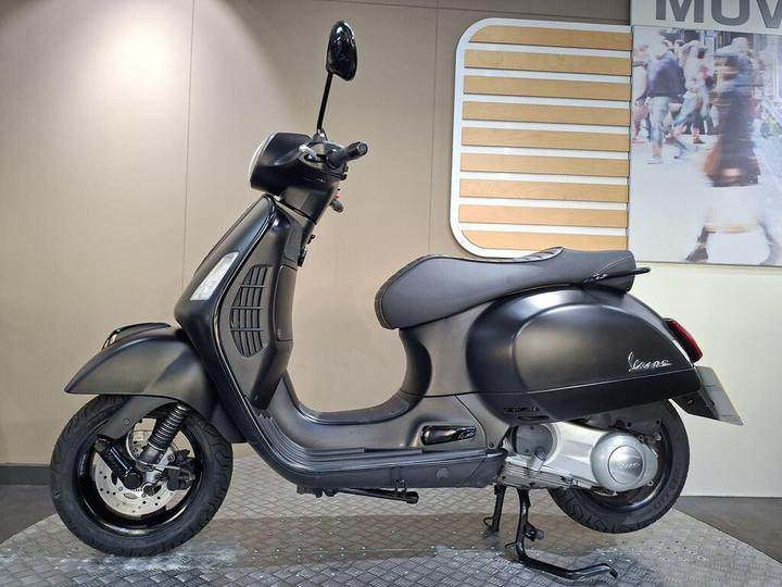A sleek black Vespa scooter viewed from the side, showcasing its curved design and chrome accents, positioned on a textured gray surface against a minimalist background.