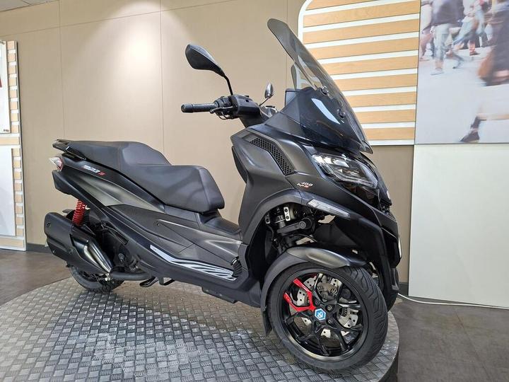 A sleek black scooter with a large windshield, displayed on a circular platform against a minimalist background. The scooter features modern design elements, including a sporty front, red suspension, and stylish wheels.