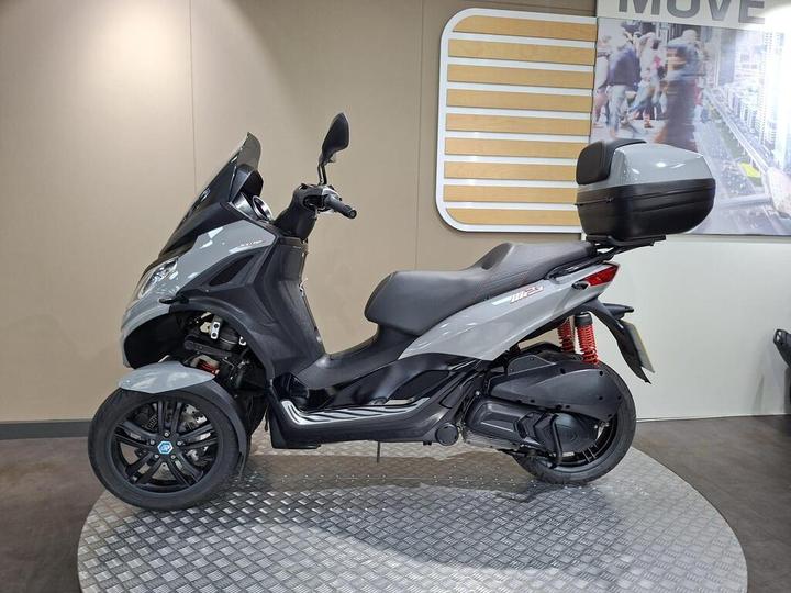 A gray three-wheeled scooter is displayed on a circular metal platform against a neutral-colored wall with a graphic design in the background. The scooter features a sleek body, a top case, and highlights like red shock absorbers.