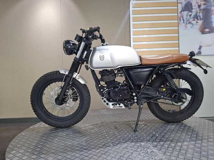 A custom motorcycle with a silver tank, brown leather seat, and black frame, displayed on a round, textured platform against a neutral background. The bike features a vintage style with fat tires and a simple headlight.