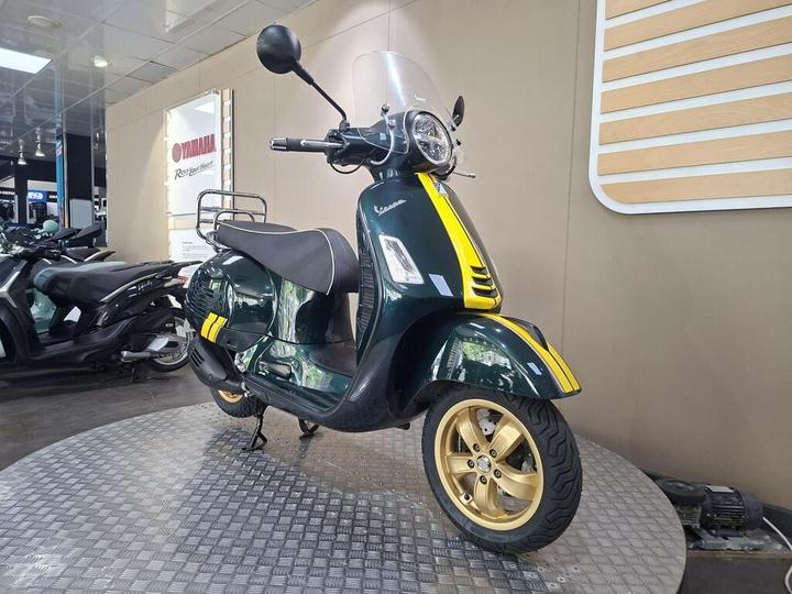 A green and yellow Vespa scooter displayed on a circular metal platform in a showroom, with other scooters visible in the background.