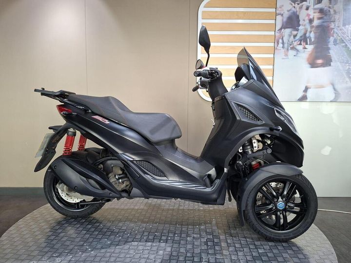Piaggio MP3 300 Sport 2022 A black three-wheeled scooter is displayed on a textured surface, showcasing its sleek design and red shock absorbers. The background features a simple, neutral wall that enhances the scooter's profile.