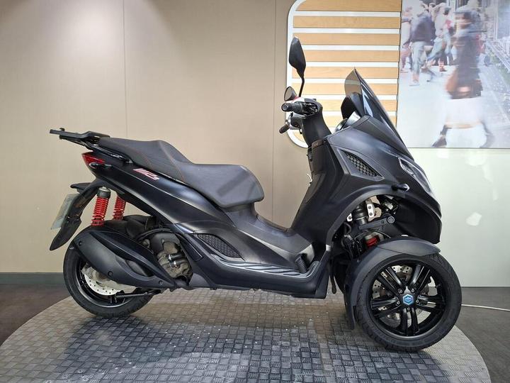 Piaggio MP3 300 Sport 2022 A black three-wheeled scooter is displayed on a textured surface, showcasing its sleek design and red shock absorbers. The background features a simple, neutral wall that enhances the scooter's profile.