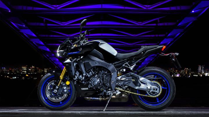 A black Yamaha MT-10 motorcycle is showcased at night, illuminated by vibrant purple lights from an overhead structure, with a city skyline visible in the background.