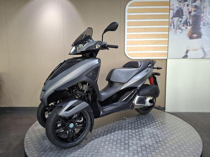 A sleek, modern three-wheeled scooter in gray and black, displayed on a circular platform with a textured surface, set against a neutral wall background.