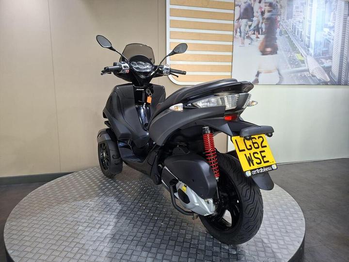A black three-wheeled scooter displayed on a circular platform, featuring a prominent rear suspension with a red spring and a visible license plate (LG62 WSE). The background shows a light-colored wall with a mural of an urban scene.