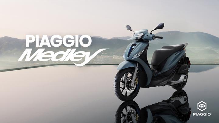 A stylish Piaggio Medley scooter in a soft blue color, positioned on a reflective surface with a serene mountainous landscape in the background. The Piaggio logo is displayed in the bottom right corner.