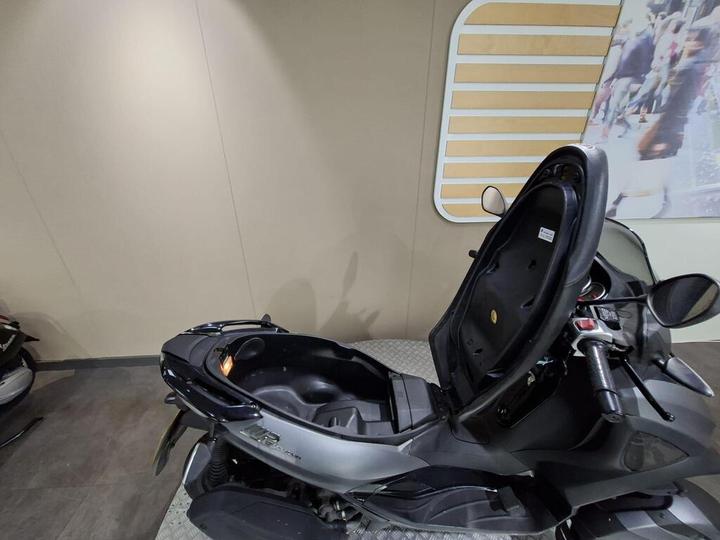 A sleek black scooter with a raised seat, revealing the storage compartment underneath, set against a neutral wall background.