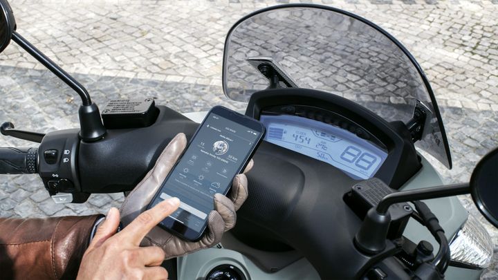 A hand wearing a glove interacts with a smartphone displaying an app, while motorcycle controls and a digital dashboard are visible in the background. The dashboard shows speed, time, and temperature readings. The scene is set on a cobblestone surface.