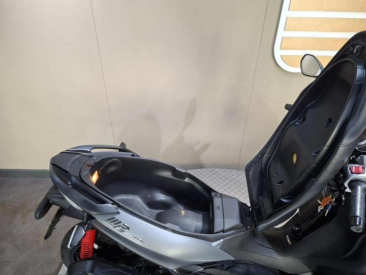 A close-up view of a scooter's seat and storage compartment, showcasing the sleek black design and visible internal space, with a light illuminating the compartment.