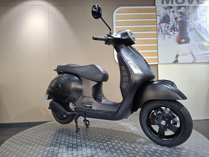 A sleek black scooter displayed on a patterned platform, featuring a rounded headlight, comfortable seat, and modern design elements, set against a softly lit wall.