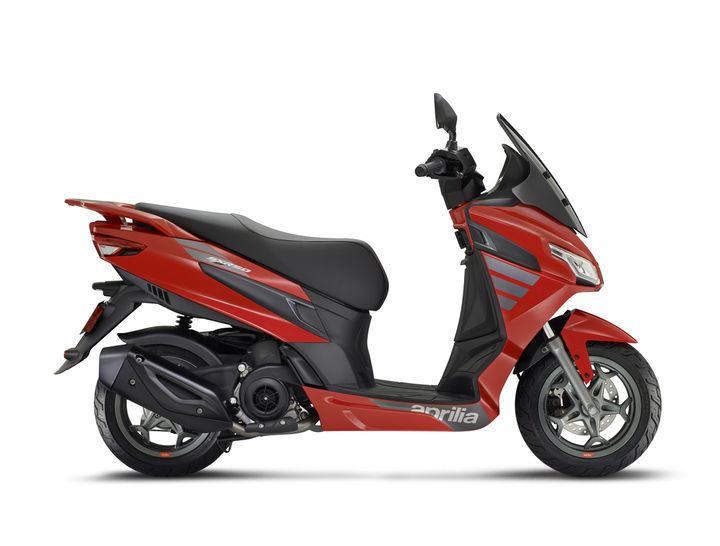 A red Aprilia scooter viewed from the side, showcasing its sleek design, prominent front windshield, and sporty features.