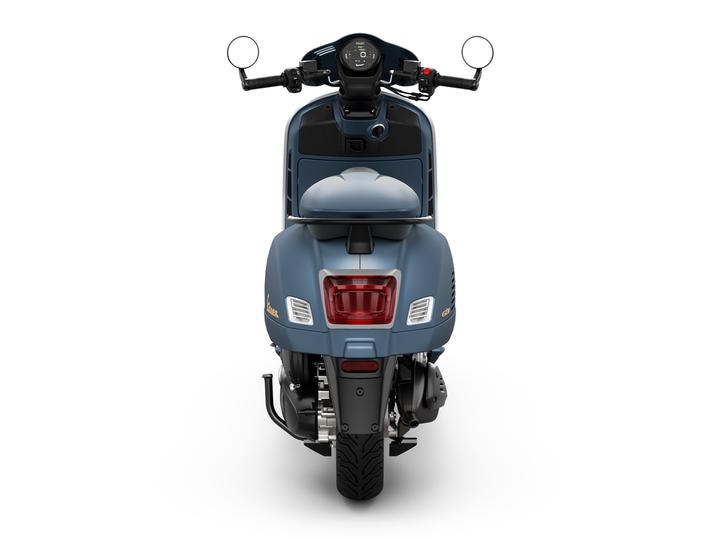 A rear view of a blue scooter with a sleek design, featuring a round rearview mirror, an analog speedometer on the handlebar, and a distinctive taillight.