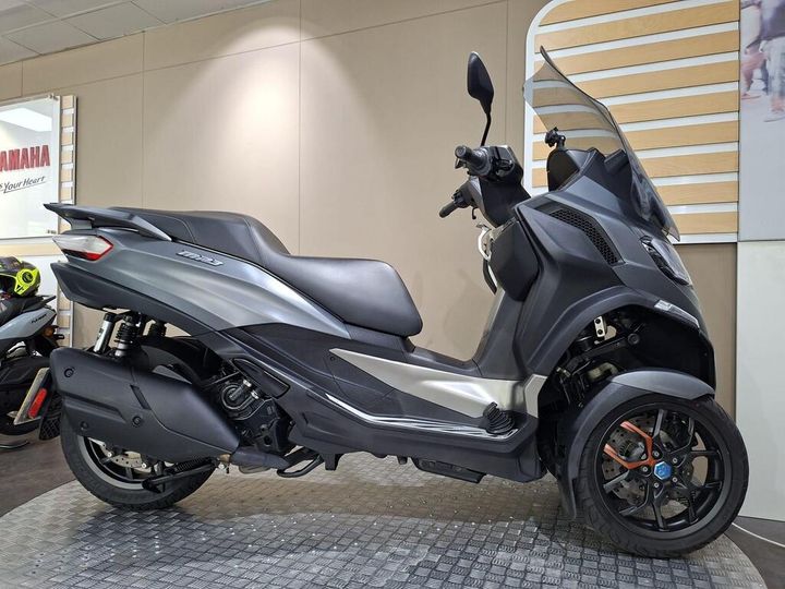 Piaggio MP3 530 HPE Exclusive 2023 A modern three-wheeled scooter in a showroom, featuring a sleek gray body, single seat, and prominent front windshield. The background includes a Yamaha sign and additional scooters.