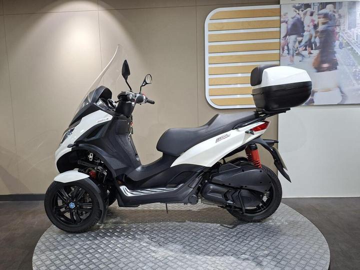 A white and black three-wheeled scooter displayed on a circular platform with a smooth background featuring a visual of people walking.