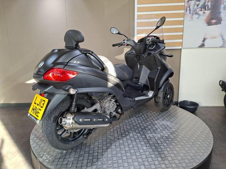 A black three-wheeled scooter displayed on a circular metal platform, featuring a rear view with a license plate reading "LJ13 HNE." The background shows a light-colored wall with blurred figures, suggesting an indoor setting.