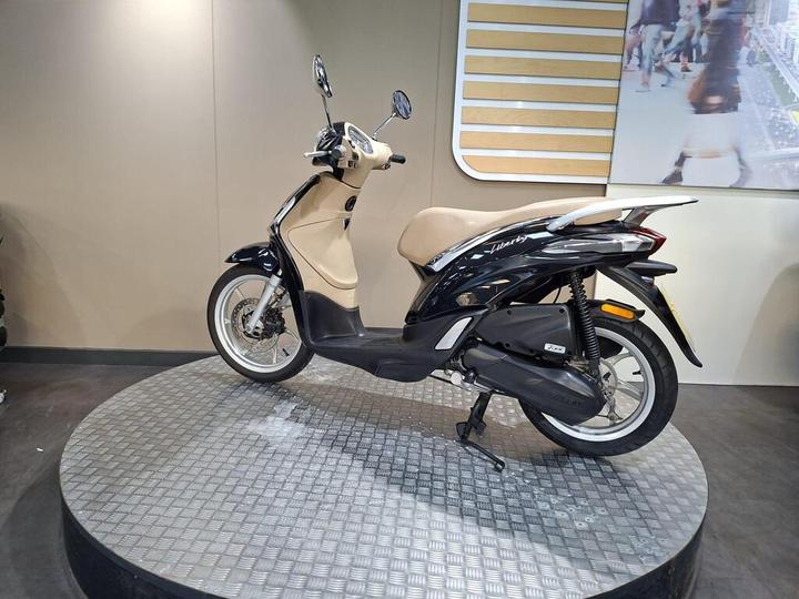 A sleek black and cream scooter displayed on a circular metallic platform, showcasing its profile with a light brown seat, mirrors, and a minimalist design against a neutral backdrop.