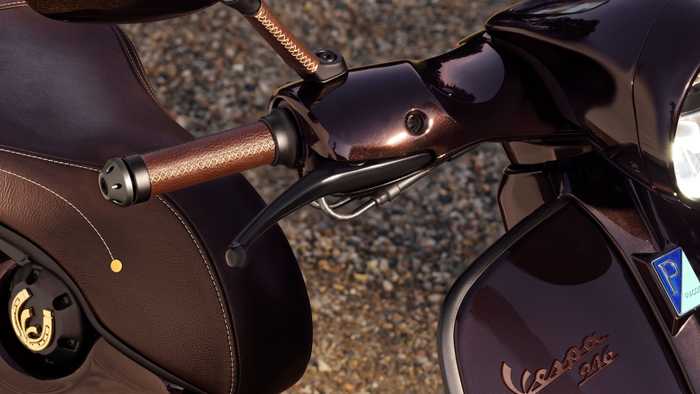 Close-up of a brown Vespa scooter's handlebar and seat, showcasing the elegant design, textured grips, and rearview mirror against a pebbled background.