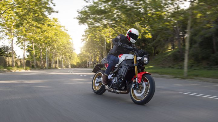 A motorcyclist in a black jacket and white helmet rides a sporty motorcycle with red and white accents on a tree-lined road, featuring blurred background motion to convey speed.
