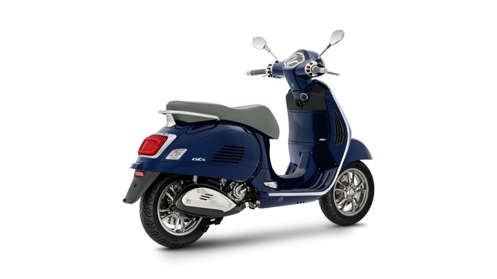 A blue Vespa scooter with a gray seat, viewed from the rear-left angle, showcasing its sleek design and chrome accents.