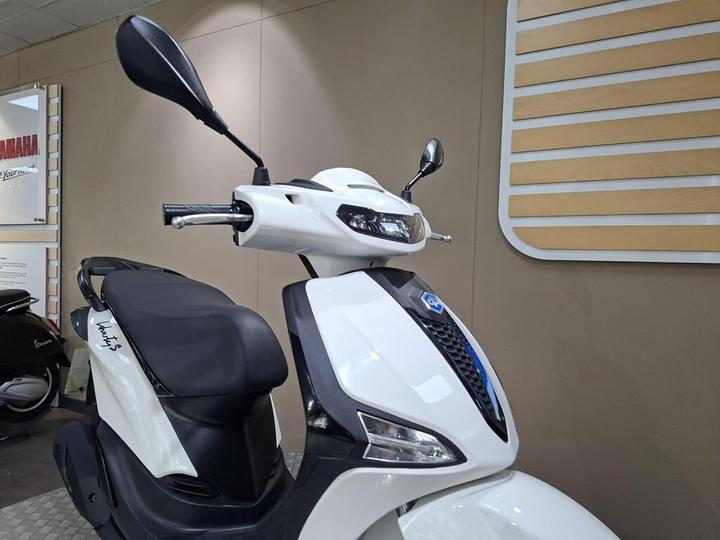 A close-up view of a white scooter featuring a sleek design, with a prominent headlight and side mirrors, displayed in a showroom backdrop.