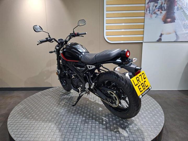 A black motorcycle displayed on a circular platform, showcasing its rear and left side, with a prominent license plate reading "LR72 EBC." The background features a simple, modern design.