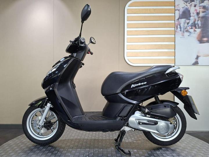 A black scooter, model Kisbee, displayed on a textured floor against a neutral wall with a wooden accent and a background image of pedestrians.