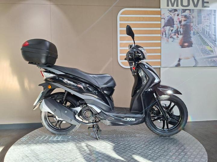 SYM Symphony ST 125 (2015) A sleek black scooter with a rear trunk stands on a textured circular platform, set against a neutral background with a partially visible urban scene.