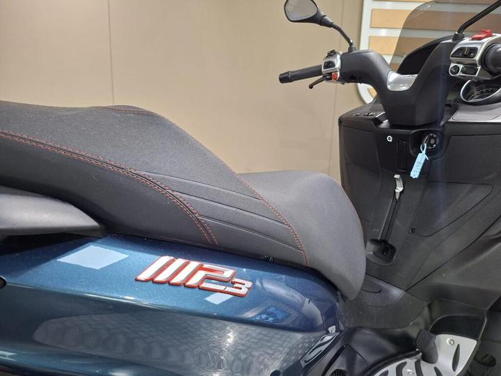 A close-up view of the seat and body of a blue scooter, featuring a prominent "MP3" logo in red, with a dashboard and handlebars visible in the background.