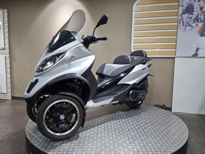A silver three-wheeled scooter showcased on a circular display platform, featuring a large windshield and modern design elements. The background includes a plain wall with a striped panel and a partially visible image of people walking.