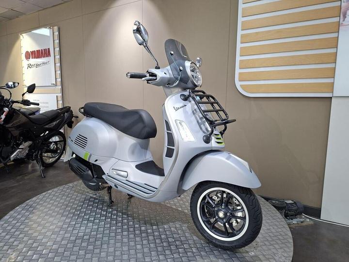 A sleek gray scooter parked on a textured display surface, featuring a prominent front with round headlights and a chrome finish, alongside a black motorcycle visible in the background. Wall signage from Yamaha is partially visible.