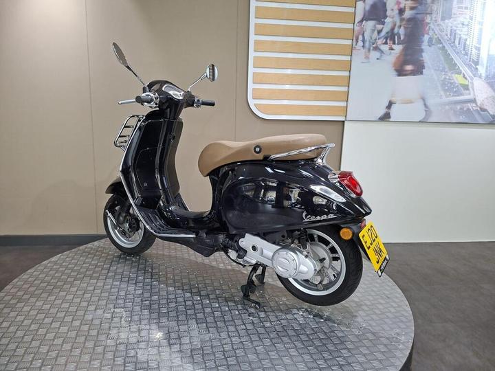 A black Vespa scooter with a brown seat is displayed on a metallic circular platform, set against a simple wall background and a digital screen showing a city scene.