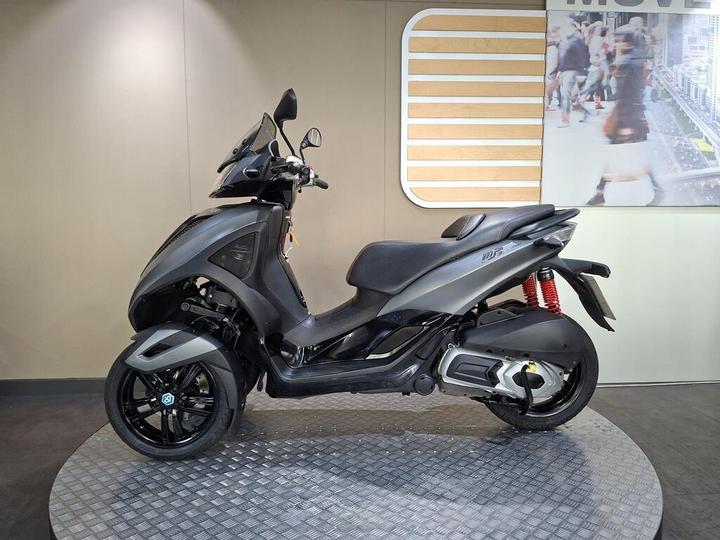 A sleek, modern scooter in matte black finish displayed on a circular metal platform, with a minimalist background featuring a blurred image of people.