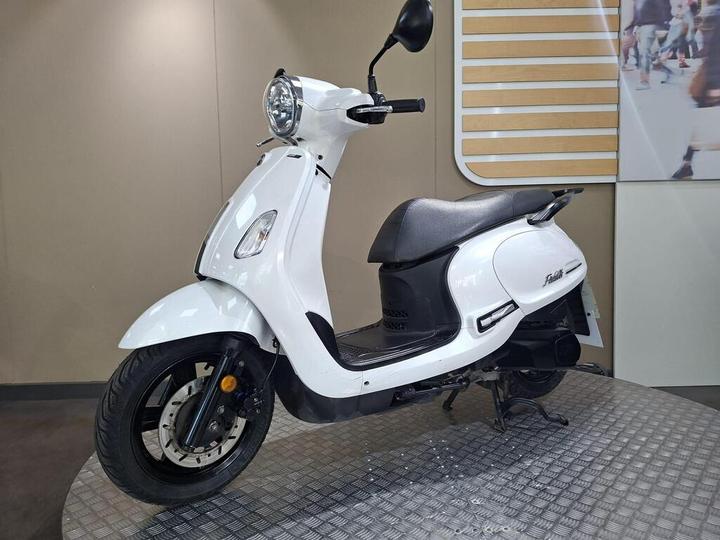 A white scooter with a rounded front, black seat, and chrome headlight displayed on a textured platform against a neutral background.