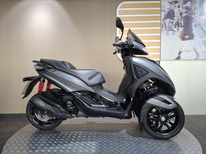 Piaggio MP3 300 (2012) A sleek, gray three-wheeled scooter displayed on a textured platform, featuring a comfortable seat, sporty design elements, and prominent rear suspension in red. The background includes a stylized wooden panel and a blurred image of people walking.