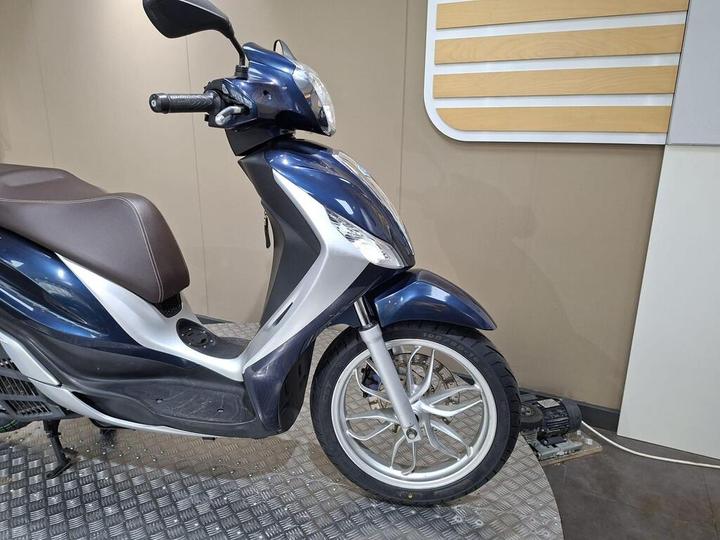 A blue and silver scooter is positioned on a metallic patterned surface, showcasing its front left side and wheel. The background features a neutral wall with horizontal wooden panels.