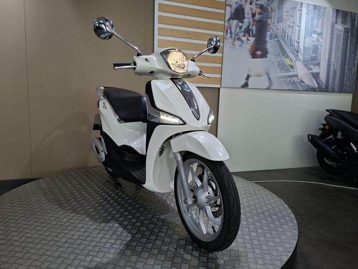 A white scooter is displayed on a circular platform in a showroom, featuring chrome mirrors and a sleek design, with a blurred background of city imagery.