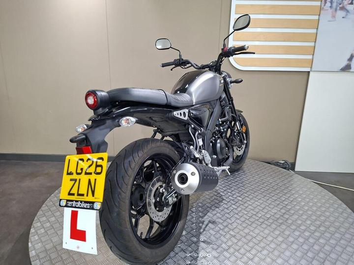 A rear view of a gray motorcycle on a circular display stand, featuring a prominent exhaust pipe and a license plate marked "LG26 ZLN." The background is a neutral-toned wall with wooden slats.