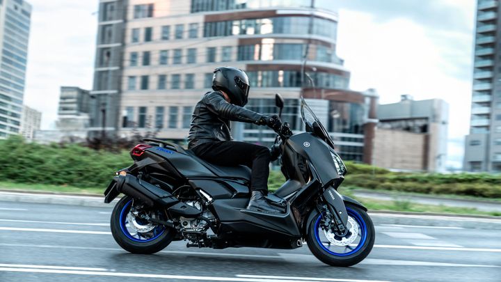 A rider in a black helmet and leather jacket is seen riding a sleek black scooter with blue accents on a city street, surrounded by modern buildings and greenery in the background.
