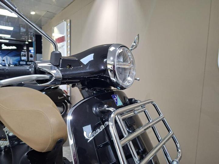 A close-up view of a black Vespa scooter featuring a chrome headlight and handlebar, with a tan seat in the foreground and a reflective interior background.