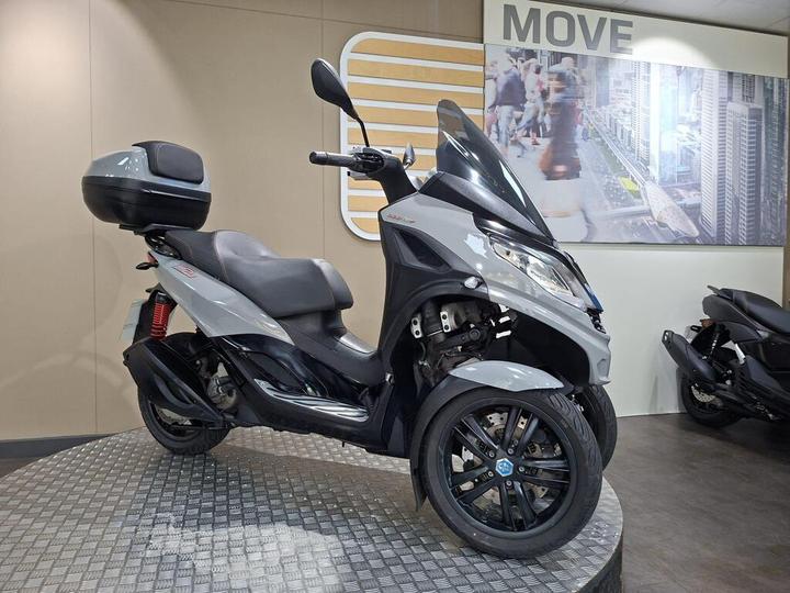 A grey scooter with three wheels parked on a circular metallic platform, displaying a modern urban setting in the background. The scooter has a prominent windshield and a top case for storage.