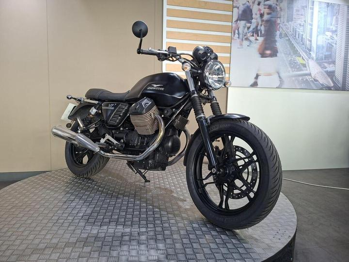 A stylish black motorcycle displayed on a circular platform, featuring a chrome exhaust and modern design elements, set against a light-colored wall and a blurred urban background.