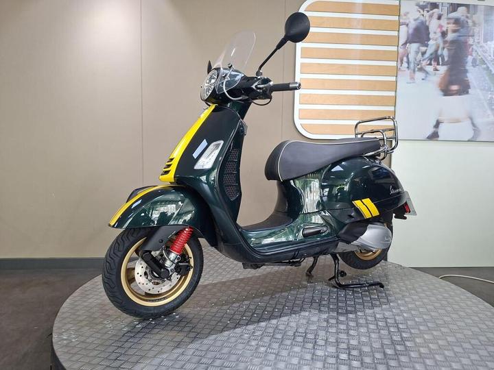A green Vespa scooter with yellow accents and a clear windscreen is displayed on a circular stand against a neutral-colored wall.