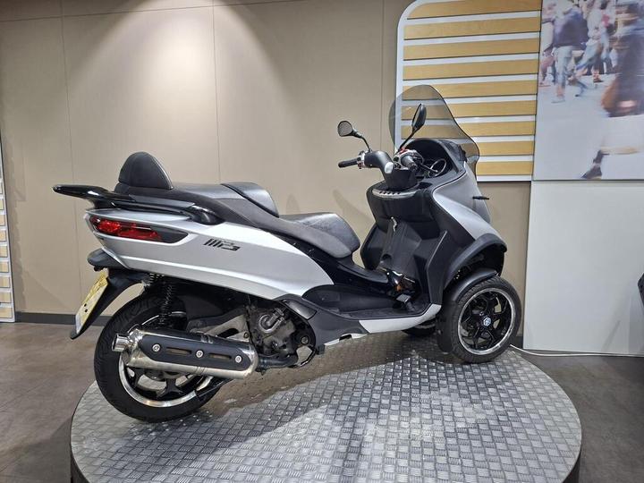 A silver and black three-wheeled scooter displayed at an exhibition, featuring a sleek design, large windshield, and a rounded rear. The scooter is positioned on a textured surface with a neutral background.