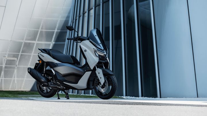 A modern scooter parked beside a sleek, contemporary building with vertical glass panels. The scooter features a predominantly white and black design, showcasing its stylish aerodynamic front.