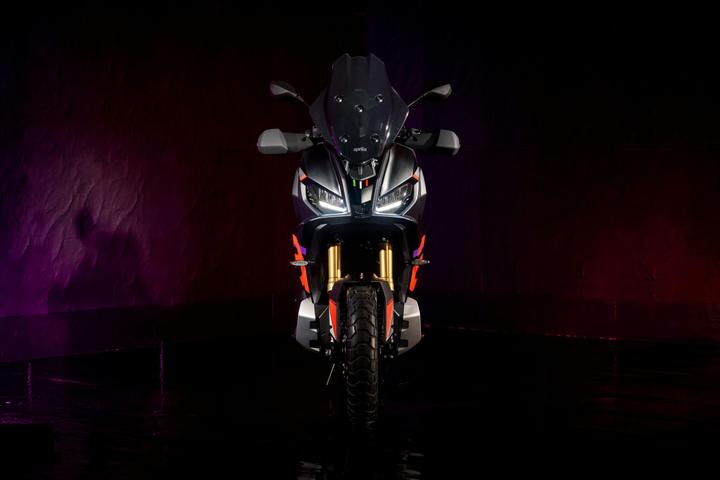 A sleek modern motorcycle with a distinctive design, featuring a matte black body, prominent front headlights, and striking orange accents, set against a dark background with colorful lighting.