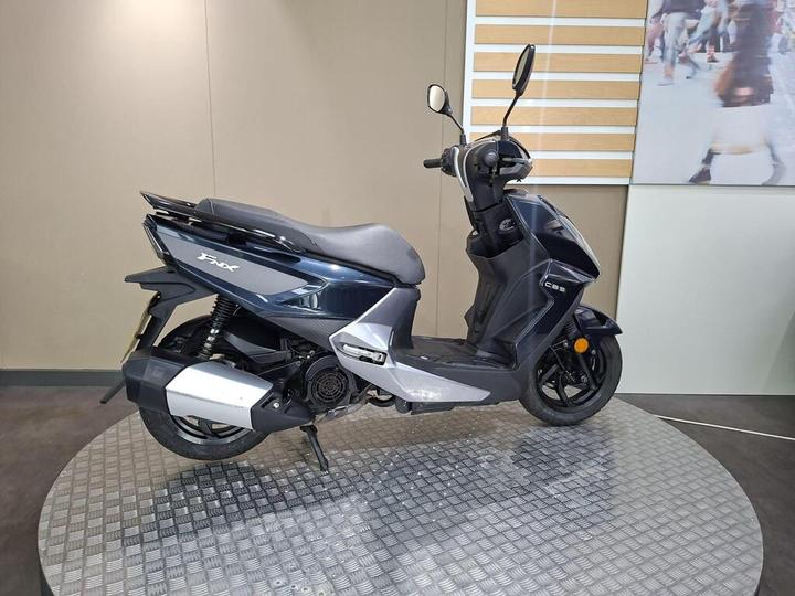 A blue and silver scooter positioned on a circular metal platform, against a neutral background with a partially visible display featuring people walking.