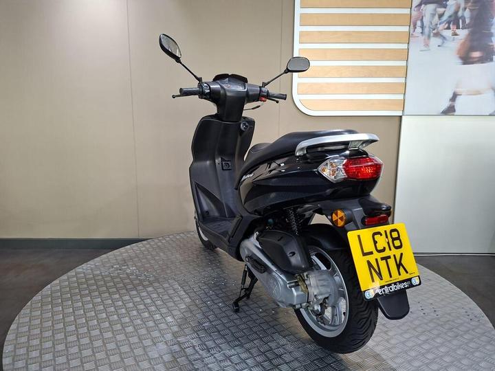 A black scooter displayed on a circular metal platform, with a visible license plate reading "LC18 NTK," set against a neutral-colored wall.