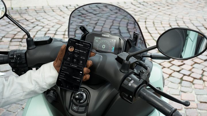 A close-up view of a motorcycle dashboard with a smartphone displaying a bike profile, featuring a Yamaha model. The image shows a gloved hand holding the phone, while various controls and a mirror are visible on the bike's handlebar. The background features cobblestone pavement.